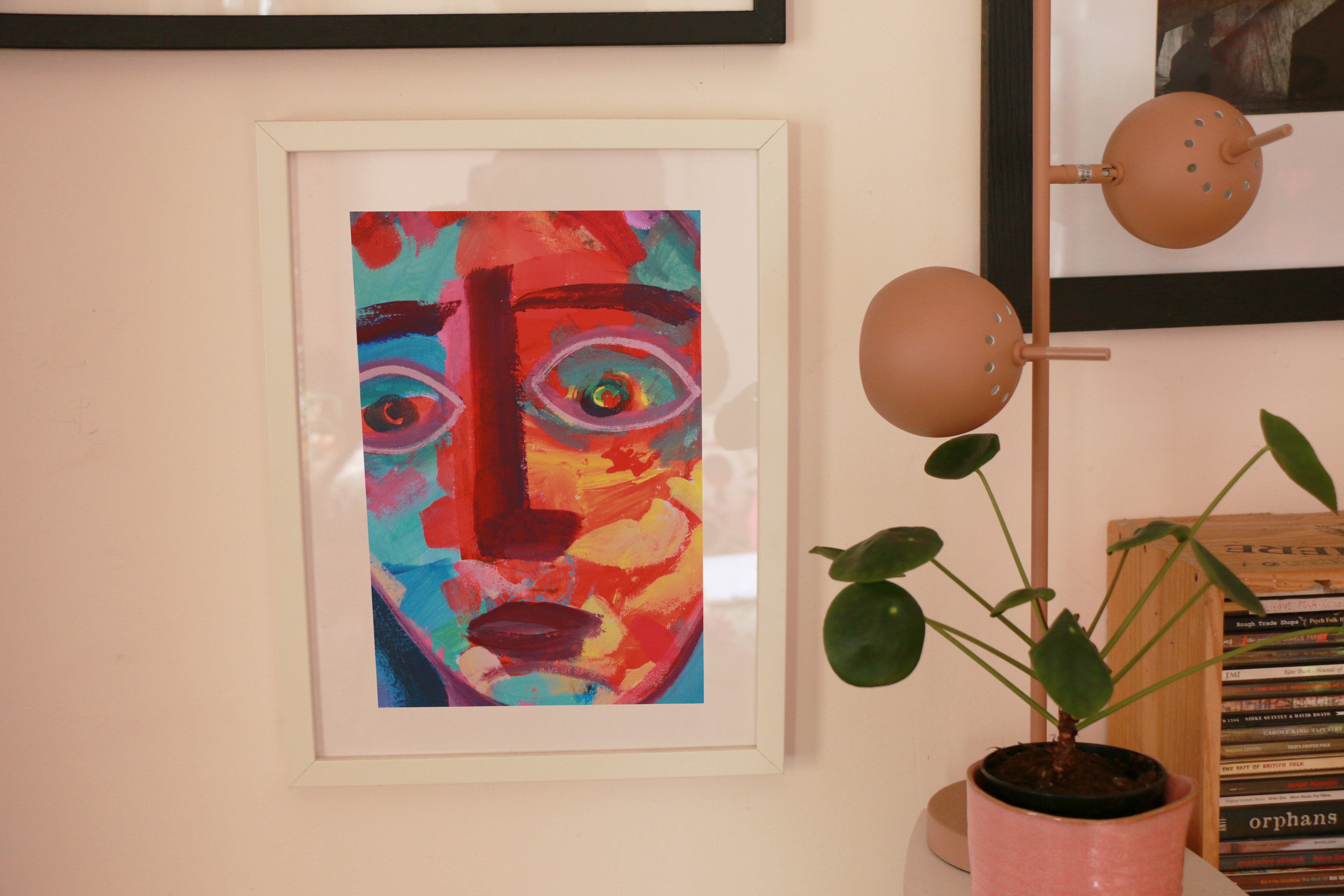 A framed picture of a colourful face on a wall, with a light and a plant beside it.